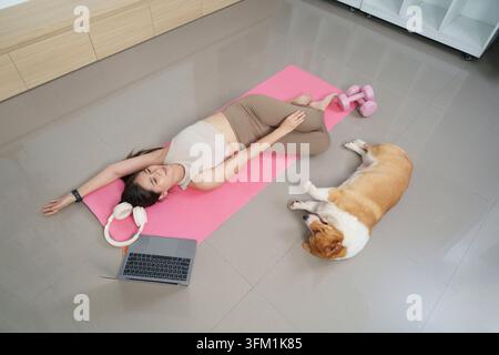 High angle shot of young Asian woman relaxing on pink exercise mat with her corgi dog, laptop and dumbbells in bright living room Stock Photo