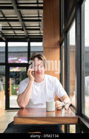 A vertical middle shot of a serious, focused Caucasian Scandinavian man in his late 30s or early 40s sit on a wooden table next to a window in a moder Stock Photo