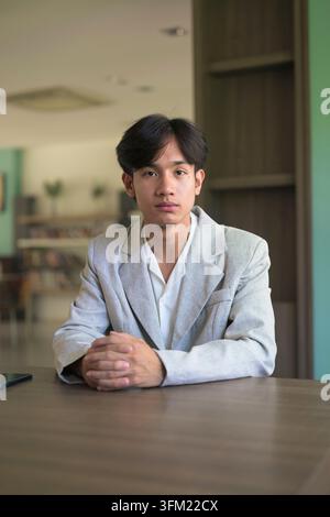 Authentic vertical lifestyle portrait of young non-binary Thai Isan professional or student sitting at a desk and looking at the camera. Dedicated han Stock Photo