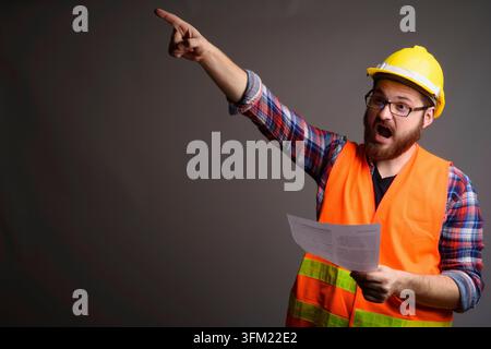 Studio portrait of a screaming bearded man in a safety vest and hard hat showing aggressively while holding a paper. Ideal for work load, construction Stock Photo