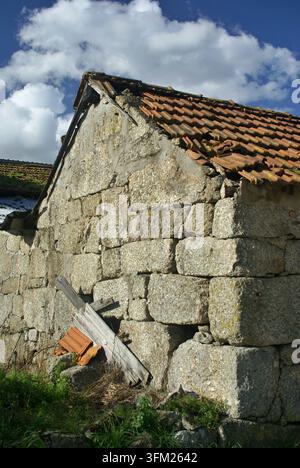 Weathered stone house wall with a broken tile roof under a blue sky Stock Photo