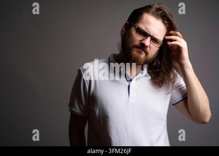 Studio portrait of a confused Caucasian man with beard and glasses scratching his head with a confused expression. Ideal for themes of difficulty, thi Stock Photo