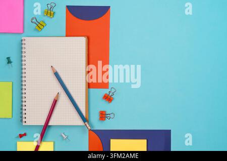 Creative flat lay of school supplies featuring an open grid notebook, two pencils, sticky notes, binder clips, and colorful geometric paper shapes on Stock Photo