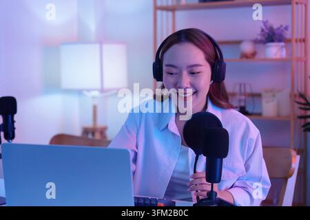 A happy young Asian female content creator wearing headphones and speaking into a microphone while recording a podcast Stock Photo