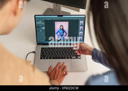 Over-the-shoulder view of two colleagues working together on a laptop, editing a portrait photograph in a digital software program Stock Photo