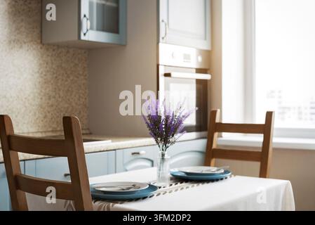 Bright modern kitchen interior decorated with wooden chairs and dining table with lavender bouquet. Cozy apartment room with natural window light and Stock Photo