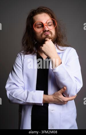 Studio portrait of a bearded young man with long hair wearing a white lab coat and oversized orange glasses, looking away thoughtfully while holding h Stock Photo