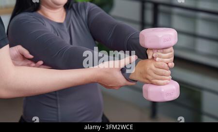 An Asian woman performs weight training with a pink dumbbell, guided by a male personal trainer at home. This image highlights fitness, exercise, and Stock Photo