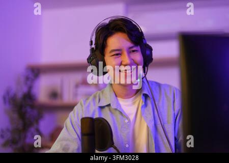 Happy Asian young man wearing gaming headset smiling while live streaming video games on computer desk in room with purple ambient lighting Stock Photo