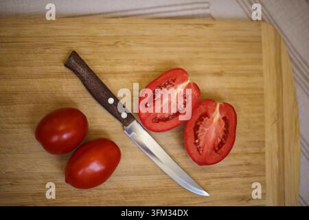 A flat lay top-down view of fresh whole and halved red plum tomatoes next to a small kitchen knife on a rustic wooden cutting board, providing generou Stock Photo