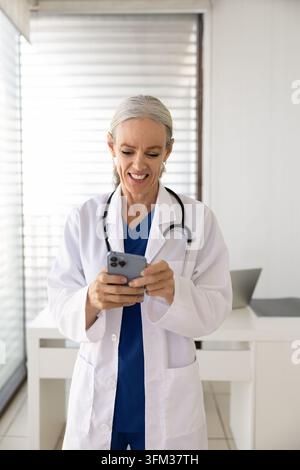 Middle-age woman doctor standing in medical office, checking smartphone Stock Photo