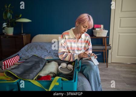 Focused woman writing packing checklist in notepad while preparing suitcase for solo travel. Stock Photo