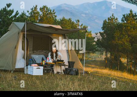 Woman cooking on a portable stove in front of a camping tent among pine trees at sunset, Ta Xua, Son La, Vietnam Stock Photo