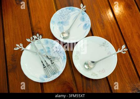 A three white porcelain plate with silver spoon and fork on polished wooden table, arranged in rustic dining setting with delicate floral decoration a Stock Photo