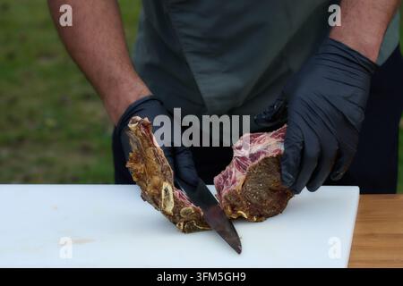Chef's gloved hands meticulously preparing a raw beef ribeye steak and bone, separating meat from bone on a white cutting board, showcasing food prepa Stock Photo