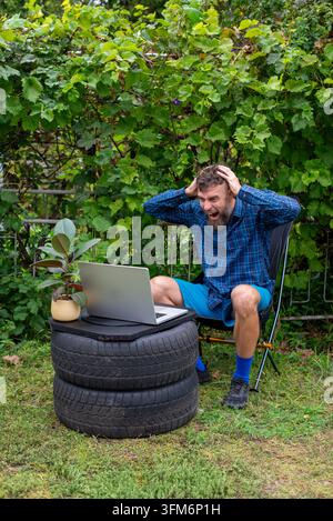 Bearded man grabs his head in shock while using a laptop on stacked tires in a backyard makeshift workspace with a potted plant. Stock Photo