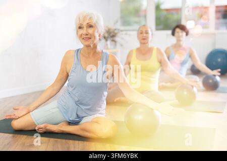Smiling mature woman doing half lotus pose with soft-ball under arm during fitness classes Stock Photo