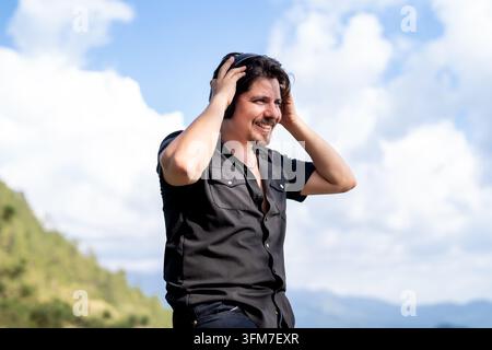 Smiling Young Man in Black Shirt Adjusting Over-Ear Headphones Outdoors with Cloudy Blue Sky and Mountain Background Stock Photo