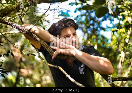 A handsome, long-haired young man in a black shirt, looking pensive as he rests his arms on a tree branch in the forest. Stock Photo