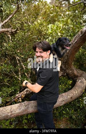 Smiling Young Man in Black Carrying His Black Mixed-Breed Dog on His Back Leaning on Fallen Tree Trunk in Forest Stock Photo