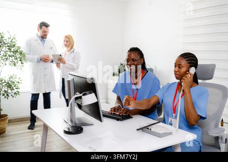 Two healthcare workers are at the computer while another staff member talks with a doctor. They are in a modern office. Stock Photo