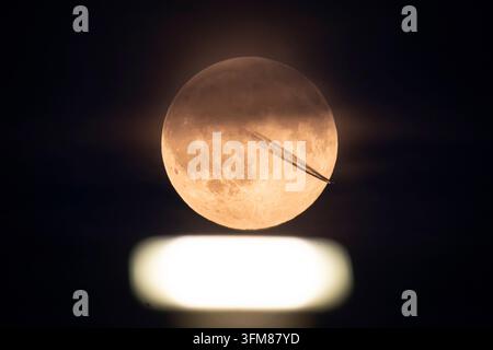 The full moon glows over a spotlight on the runway of Frankfurt Airport, while the contrails of a passing aircraft are emerging in front of the Earth' Stock Photo