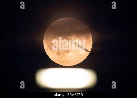 The full moon glows over a spotlight on the runway of Frankfurt Airport, while the contrails of a passing aircraft are emerging in front of the Earth' Stock Photo