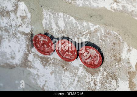 Three red plaster covers on electrical installation boxes embedded in a rough plastered wall showing construction details during interior renovation w Stock Photo