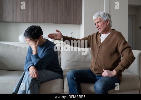 Manipulative narcissistic elderly husband blaming and shaming aged woman sitting on couch at home. Stock Photo