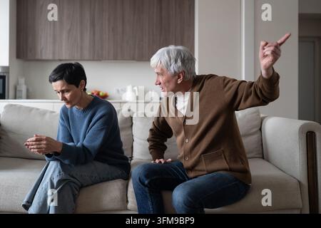 Furious senior husband talking angry yelling at sad distressed wife while couple sitting on couch. Stock Photo