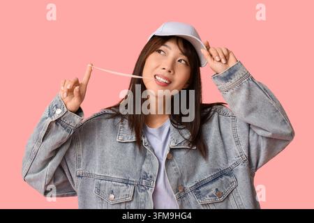 Youth culture and carefree lifestyle, young woman chewing bubble gum and stretching it with fingers wearing denim jacket and baseball cap on pink back Stock Photo