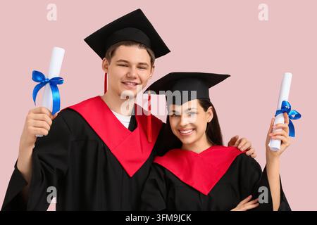 Higher education graduation and academic success, happy young students in caps and gowns holding diplomas on a pink background Stock Photo