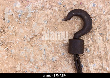Rusty iron lifting hook attached to a thick chain lying on rough gravelly soil in an overhead view. Stock Photo