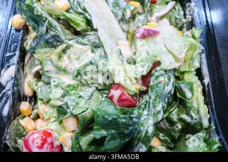 Prepared mixed green salad with halved cherry tomatoes, sweet corn kernels, and creamy dressing in black plastic takeaway food tray. Stock Photo