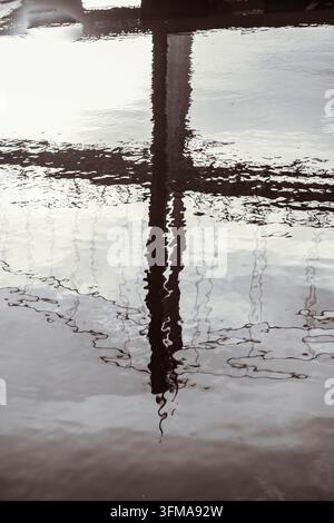 Serene water reflects a tall structure, sending quiet ripples across its surface. Reflections sway gently, mirroring stillness and motion. Monochrome tones enhance tranquility Stock Photo