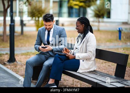 Diverse business professionals sharing ideas and using digital devices while sitting on a park bench Stock Photo