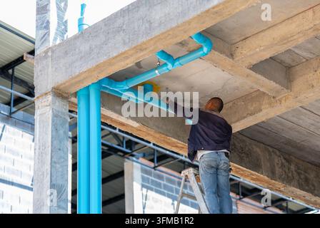 Plumber on a stepladder installing blue PVC water supply pipes beneath a concrete building frame on a construction site. Stock Photo
