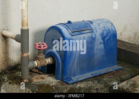 A bright blue protective cover shields a water meter, attached to a brass valve and grey pipes. The scene is set outdoors on a rough concrete surface. Stock Photo