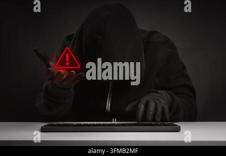 Anonymous scammer hooded cybercriminal holds glowing warning symbol while using keyboard. Financial cyber fraud, online scam risk, digital threats and Stock Photo