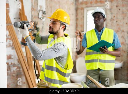 Portrait of man builder working with handheld demolition hammer at indoor construction site, his chef standing behind and making notes on clipboard Stock Photo