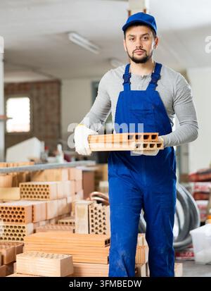 Portrait of focused young man worker in uniform carrying red bricks at indoor building construction site Stock Photo