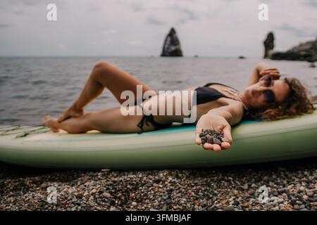 Small pebbles held out by young woman relaxing on paddleboard near rocky ocean shore Stock Photo