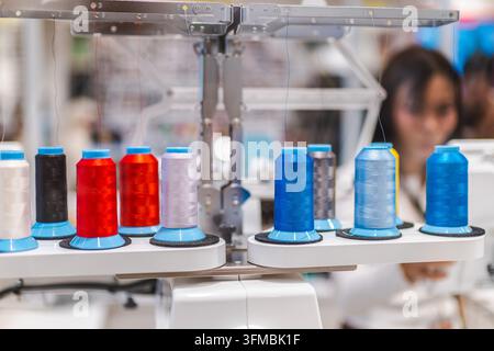 Colorful thread spools sit on industrial textile machinery while a worker is blurred in the background. Stock Photo