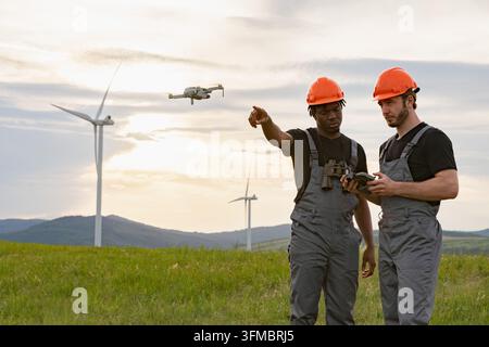 Two engineers in hard hats operating a drone near wind turbines at sunset Stock Photo
