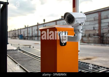 White dome security camera mounted on orange access control post overlooking industrial loading dock area at dusk under overcast cloudy sky Stock Photo