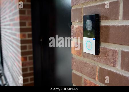 Smart doorbell camera mounted on red brick exterior wall beside residential front door in daytime, showing illuminated ring button, lens, and security Stock Photo