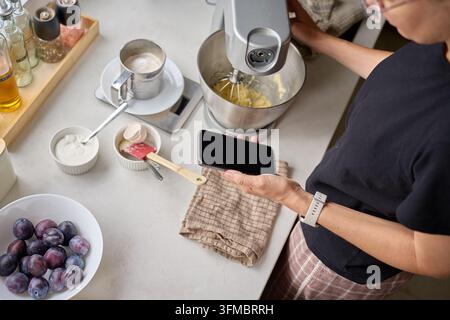 Woman holding a smartphone beside a stand mixer and baking tools on the kitchen counter. Home baking, cooking and using a phone while preparing food Stock Photo