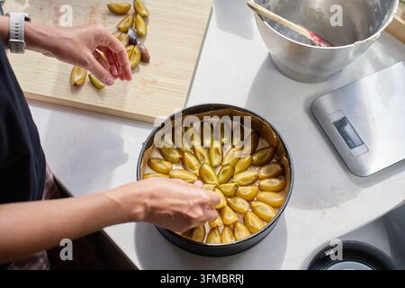 Woman arranging sliced plums on dough while making pie in kitchen. Person adding fruit to round baking pan beside mixing bowl and kitchen scale. Home Stock Photo