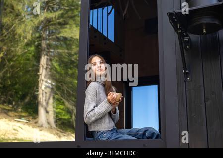 Young woman sitting by an open window holding a warm drink, peaceful lifestyle portrait in a modern forest house with lush green nature outside. Stock Photo