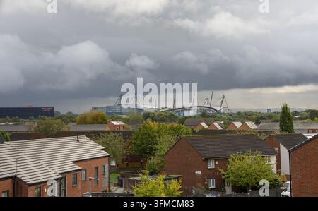 Manchester's Etihad Stadium rising behind suburban brick houses and green trees beneath dramatic, dark storm clouds in Greater Manchester, UK. Stock Photo